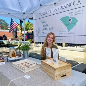 May include: A woman stands behind a table with a sign that reads "Pendleton Granola" and "Made Locally in Pendleton, SC". The sign also lists the ingredients: oats, honey, dried fruit, and nuts. The woman is holding two bags of granola. The table is covered in a white tablecloth and has a wooden box and a tray of granola on it.