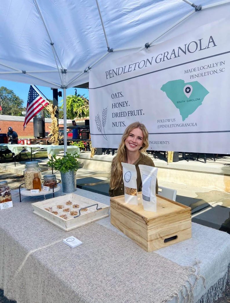 May include: A woman stands behind a table with a sign that reads "Pendleton Granola" and "Made Locally in Pendleton, SC". The sign also lists the ingredients: oats, honey, dried fruit, and nuts. The woman is holding two bags of granola. The table is covered in a white tablecloth and has a wooden box and a tray of granola on it.