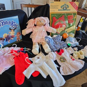 May include: A collection of vintage Cabbage Patch Kids items, including a doll with pink yarn hair, various outfits, shoes, and a calendar. The doll is wearing a pink dress. Other items include a box set and a small orange ball.