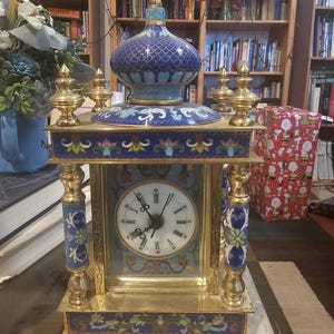 May include: A gold and blue cloisonn&eacute; clock with a white face and black Roman numeral hour markers. The clock has a decorative top and ornate details.