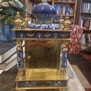 May include: A gold and blue cloisonn&eacute; clock with a decorative design. The clock has a gold base and a blue and gold top with a floral design. The clock is on a table with a book and a Christmas present in the background.