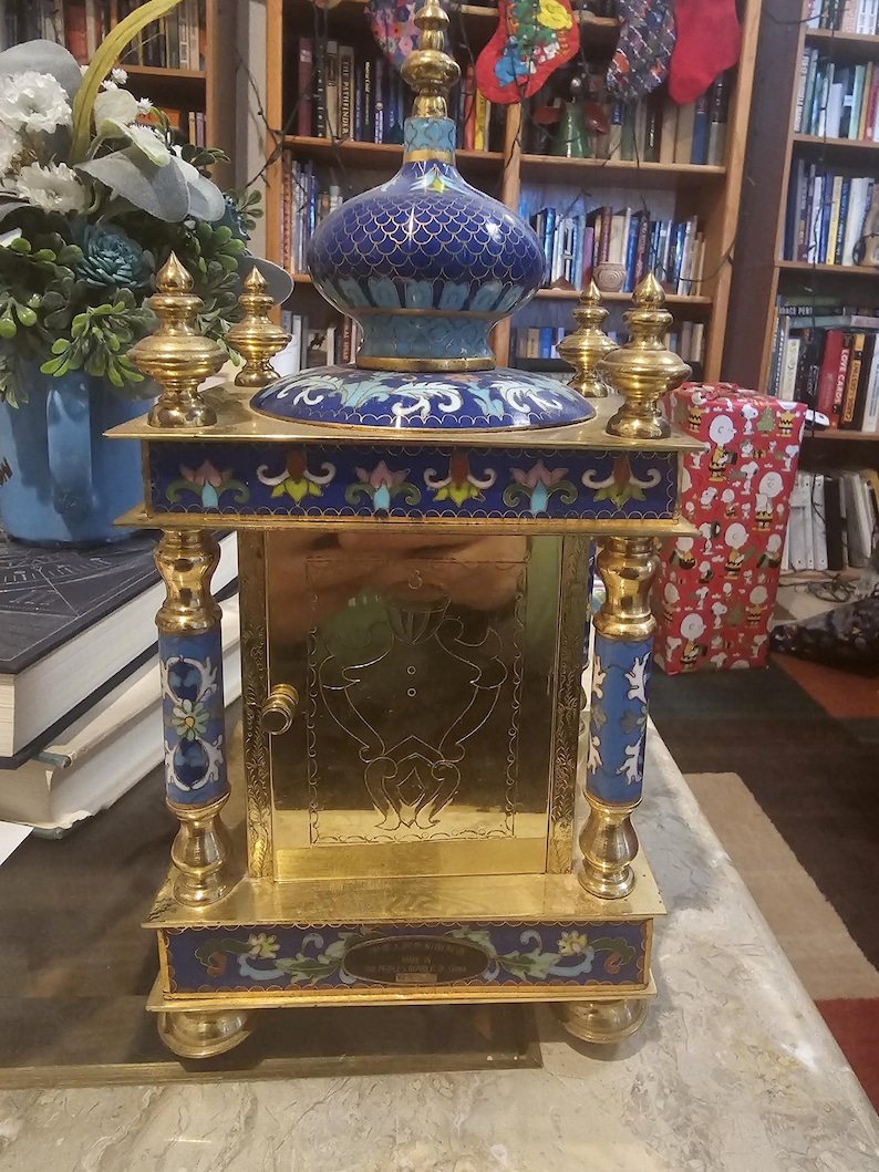 May include: A gold and blue cloisonn&eacute; clock with a decorative design. The clock has a gold base and a blue and gold top with a floral design. The clock is on a table with a book and a Christmas present in the background.