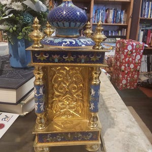 May include: A gold and blue cloisonn&eacute; clock with intricate floral designs and a gold base. The clock is on a table with a book and a wrapped gift in the background.