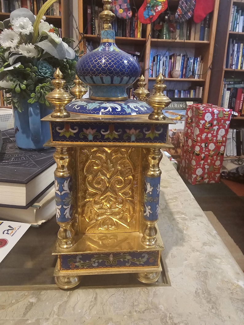 May include: A gold and blue cloisonn&eacute; clock with intricate floral designs and a gold base. The clock is on a table with a book and a wrapped gift in the background.