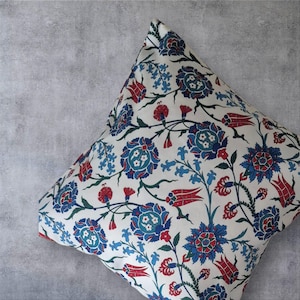 May include: Square decorative pillow with a floral pattern in red, blue, and green on a white background. The design features stylized flowers and foliage. The pillow is placed against a gray background.