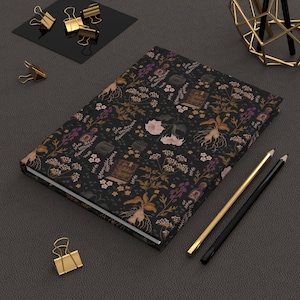 May include: A hardcover journal with a dark floral pattern featuring botanical illustrations in shades of brown, purple, and cream. The journal is surrounded by gold and black office supplies, including binder clips and pencils.