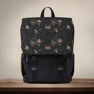 Peut inclure: Sac à dos noir avec un rabat orné d'un motif floral sombre avec des papillons de nuit, des étoiles et des croissants de lune. Le sac à dos a deux poches avant et une poignée supérieure. Les sangles sont noires avec des coutures blanches.
