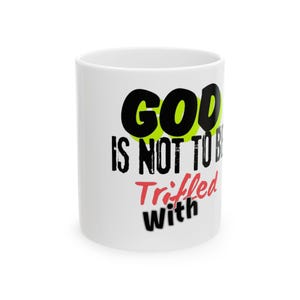 May include: White ceramic mug with the text "GOD IS NOT TO BE TRIFFLED WITH" in black, green, and red lettering. The mug is cylindrical and has a glossy finish. The text is centered on the mug.