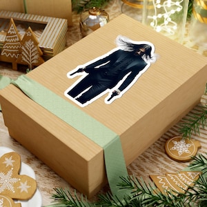May include: A sticker featuring a person in a black suit holding two guns. The sticker has a white border and is placed on a brown gift box with a green ribbon. Christmas decorations are in the background.