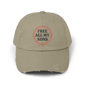 May include: A khaki-coloured baseball cap with a curved peak. The cap displays the text "FREE ALL MY SONS" within a red barbed wire design. The cap has a distressed appearance, with worn edges and a vintage aesthetic.