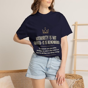 Peut inclure: Un t-shirt bleu marine à manches courtes avec une icône de couronne dorée et le texte : AUTHORITY IS NOT GRANTED-IT IS REMEMBERED. The moment you stop asking permission to exist is the moment you meet the self that was always sovereign.
