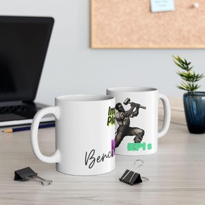 May include: Two white ceramic mugs with a black and white illustration of a person holding a hammer. The text on the mugs reads "Imagine Place" and "Bench Press".