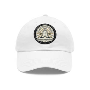 May include: White baseball cap featuring a circular patch with praying hands and the text "Praise changes You." The patch also includes the phrase "Praise does not change God." The design incorporates gold and beige accents.