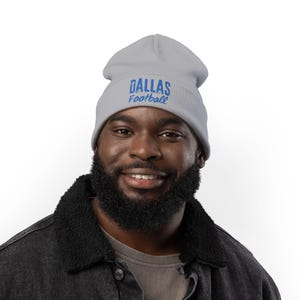 May include: A light gray beanie with the words "DALLAS Football" in blue. The beanie is made of a soft material and is designed to keep the head warm. The beanie is a great accessory for any fan.