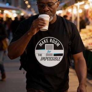 May include: A black t-shirt with the text "MAKE ROOM for THE IMPOSSIBLE" in a circular design. The person in the image is wearing a black beanie, glasses, and brown pants, and is holding a coffee cup.