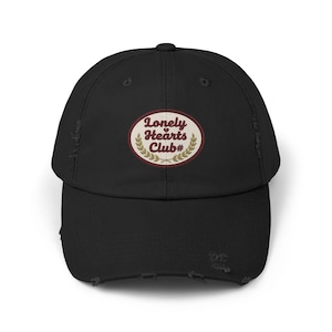 May include: Black distressed baseball cap with a curved brim. The cap features an oval patch with the text "Lonely Hearts Club#" in a serif font, surrounded by a laurel wreath design. The cap has a worn, vintage look.