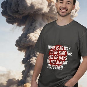 May include: Dark gray t-shirt with the text "THERE IS NO WAY TO BE SURE THE END OF DAYS HAS NOT ALREADY HAPPENED" in white and red. A man smiles in front of a large mushroom cloud.
