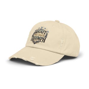 May include: A beige baseball cap with a distressed, frayed brim and front panel. The cap features a dark blue and gold graphic print with the text "SHOUT with a Voice of TRIUMPH" surrounded by small bird silhouettes and decorative line work.