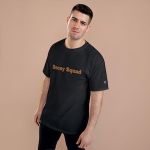 Puede incluir: Camiseta negra con el texto "Decoy Squad" en naranja. La camiseta tiene mangas cortas y cuello redondo. El logo de Champion es visible en la manga izquierda.