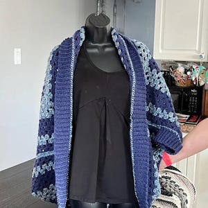 May include: A blue and light blue crocheted cardigan with a shawl collar. The cardigan is open at the front and is being worn over a black vest top.