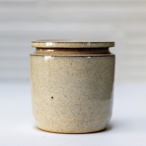 May include: A small, cream-colored ceramic jar with a lid. The jar has a speckled texture and a slightly darker brown rim.