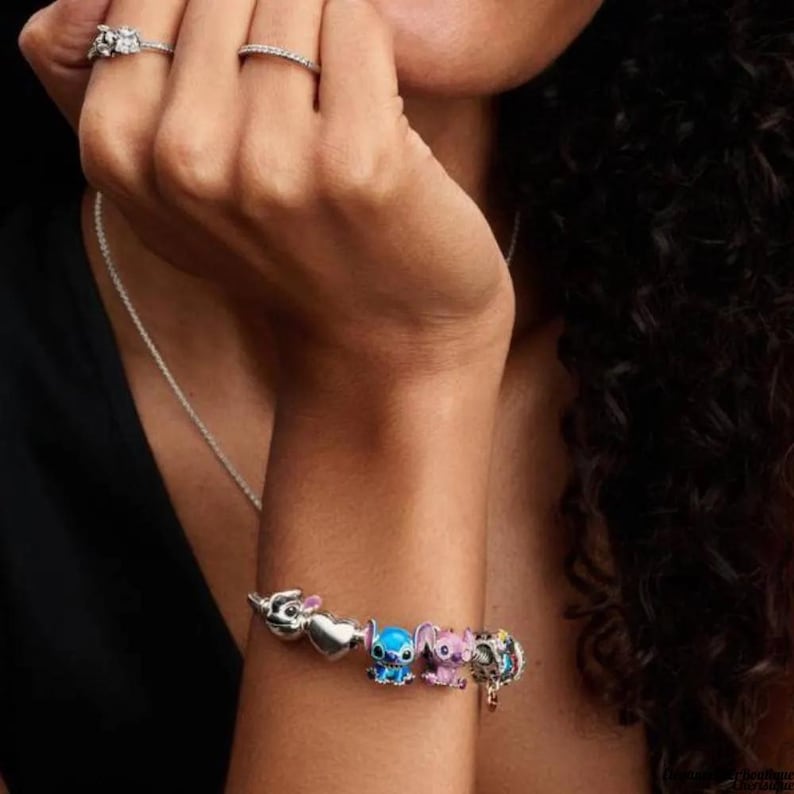 May include: A silver charm bracelet featuring Disney characters. The bracelet includes charms of Stitch, Angel, and other characters. The bracelet is worn on a person's arm, with rings on the fingers.