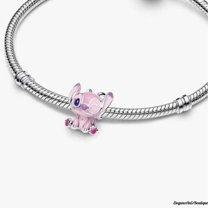 May include: A pink and purple charm featuring a cartoon character, attached to a silver bracelet. The charm is a small, detailed figure with large ears and a winking expression. The bracelet has a textured, snake-like design.