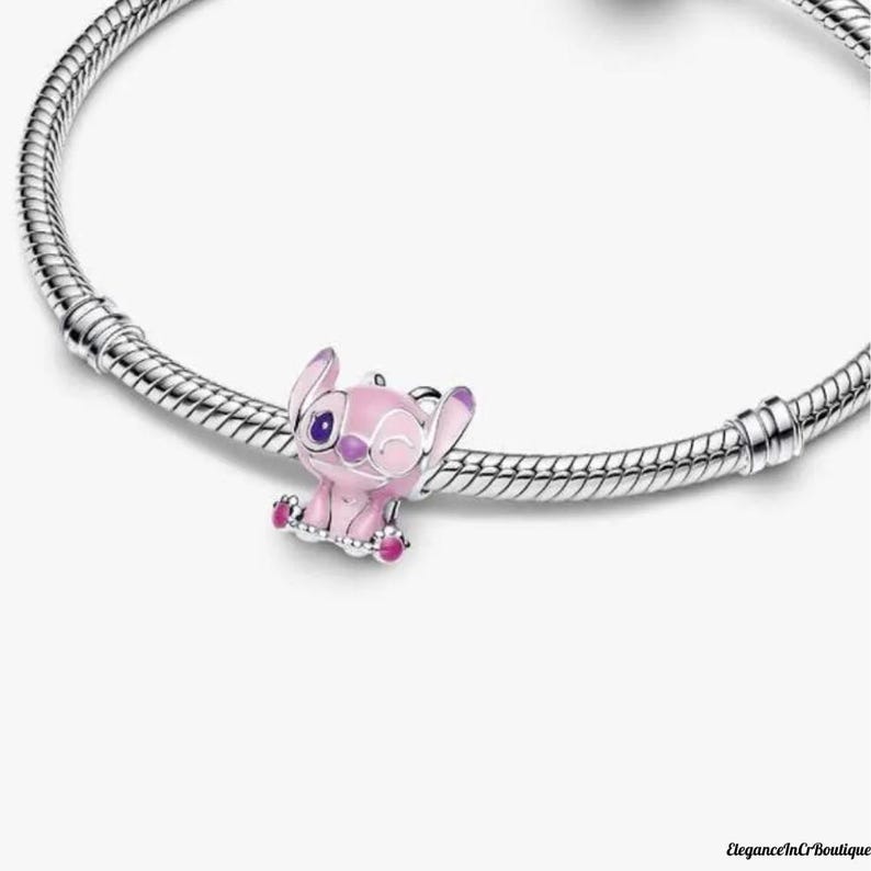 May include: A pink and purple charm featuring a cartoon character, attached to a silver bracelet. The charm is a small, detailed figure with large ears and a winking expression. The bracelet has a textured, snake-like design.