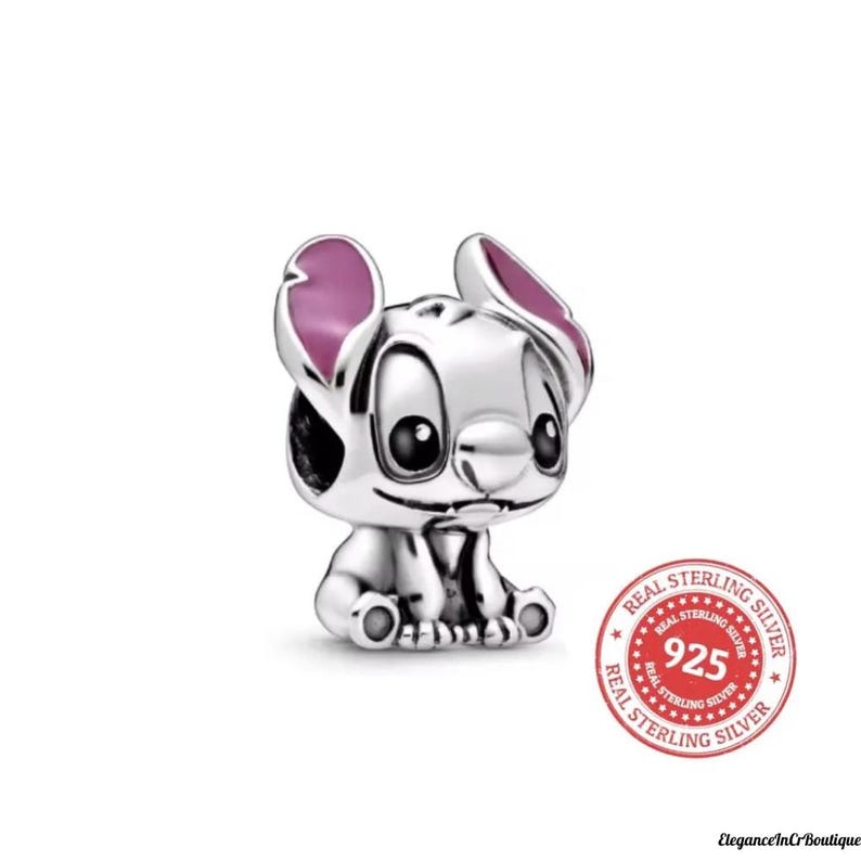 May include: A silver-colored charm featuring a cartoon character with large black eyes and pink-colored ears. The charm is in a sitting position and has a small smile. The charm has a "925 Real Sterling Silver" stamp.