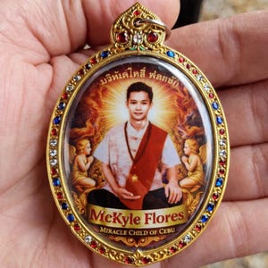 Puede incluir: Colgante dorado ovalado con un retrato de un hombre con camisa blanca y faja roja, rodeado de ángeles. El colgante tiene un borde decorativo con piedras rojas, azules y transparentes. Se ve el texto "McKyle Flores" y "MIRACLE CHILD OF CEBU".
