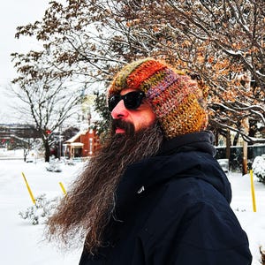 May include: A chunky knit beanie featuring a variegated color palette of orange, brown, green, and purple yarn. The hat has a thick, ribbed texture and is worn with black sunglasses, a dark hooded jacket, and a long, full brown beard.