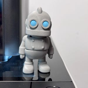 May include: A gray robot figurine with bright blue eyes. The toy robot has a rounded head, a cylindrical body, and articulated arms and legs. The robot is standing on a reflective black surface.