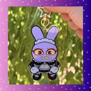 May include: A clear acrylic keyring featuring a cartoon bunny in a maid outfit. The purple bunny has large red eyes and a worried expression. The keyring has a gold-coloured star-shaped clasp.