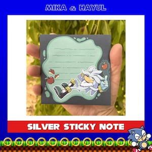 May include: A square sticky note with a dark gray border and a light green central area featuring horizontal lines for writing. The design includes a cartoon illustration of a silver hedgehog character, red apples, and apple slices.