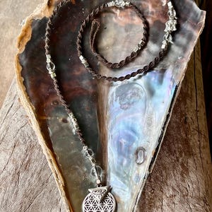 May include: A brown cord necklace with clear crystal beads and a silver pendant. The pendant features a geometric design with an eye symbol. The necklace is displayed on a pearlescent surface.