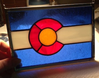Pode incluir: Uma janela de vitral representando a bandeira do estado do Colorado. A bandeira apresenta um círculo vermelho grande com um círculo amarelo menor no centro, rodeado por um fundo branco. O círculo está colocado sobre um fundo azul com uma faixa branca.