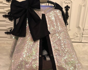 May include: A white iridescent sequin car seat cover with a black bow on top. The cover has a black trim and a tag that says "Covered In Love".