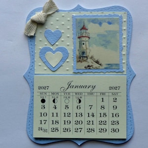 May include: A handmade January 2027 calendar with a blue border and a lighthouse illustration. Features a white bow, two blue hearts, and a calendar grid. The calendar is a decorative item.