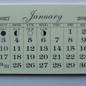 May include: A small, rectangular calendar for January 2027, printed in black ink on a light green card. The calendar grid includes the days of the week, dates, and moon phases. The word "January" is written in an elegant script.