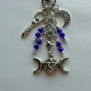 May include: A silver-toned charm cluster featuring a crescent moon, a stylized female figure, and a triple moon pendant with a pentagram. Strands of blue beads and a star charm add detail to this mystical accessory.