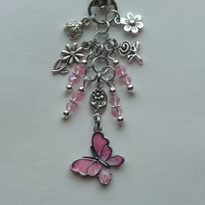 May include: Silver-toned keychain with multiple charms, including flowers, leaves, and pink beads. A pink butterfly charm with black details hangs at the bottom. The keychain is designed with a variety of decorative elements.
