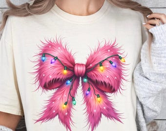May include: Cream-colored t-shirt featuring a large, vibrant pink bow design. The bow is adorned with colorful string lights, creating a festive and eye-catching look. The design is centered on the shirt, making it a statement piece.