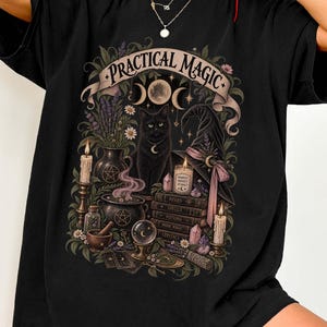 May include: A black t-shirt featuring a graphic print with the text "PRACTICAL MAGIC" in a banner. The design includes a black cat, a witch hat, a cauldron, a crystal ball, candles, books, and various botanical elements in muted earth tones.