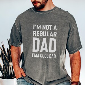 May include: Gray t-shirt with the text "I'M NOT A REGULAR DAD I'M A COOL DAD" in white block letters. The shirt has a vintage, faded look. The person is wearing blue jeans and a brown watch.