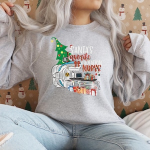 Puede incluir: Una sudadera gris con un diseño navideño que presenta una máquina de radiación, un árbol de Navidad y el texto "La enfermera de radiación favorita de Santa".