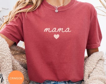 May include: Crimson-colored t-shirt with the word "mama" in a cursive font and a small heart beneath it. The shirt has short sleeves and a crew neck. The model is wearing a silver necklace.