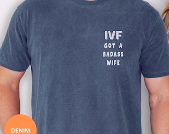 May include: A denim blue short-sleeved t-shirt featuring white text on the left chest area that reads IVF GOT A BADASS WIFE. The shirt has a crew neckline and a slightly faded, textured appearance across the fabric surface.