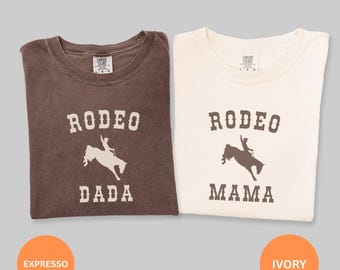 May include: Two t-shirts, one espresso-colored and one ivory-colored, each with a rodeo-themed graphic. The espresso shirt says "RODEO DADA" and the ivory shirt says "RODEO MAMA". Both shirts feature a silhouette of a rider on a bucking horse.