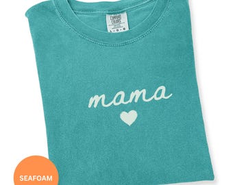 May include: Seafoam green t-shirt with the word "mama" in white script and a white heart below. The shirt is folded and has a Comfort Colors tag in the upper right corner. An orange circle with the word "SEAFOAM" is in the lower left corner.