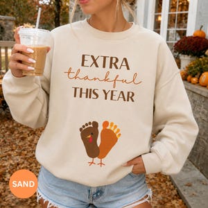 May include: A sand-colored crewneck sweatshirt featuring the brown and orange text "EXTRA thankful THIS YEAR" above a graphic of two footprints shaped like a turkey. The sweatshirt has long sleeves and a ribbed collar, cuffs, and hem.
